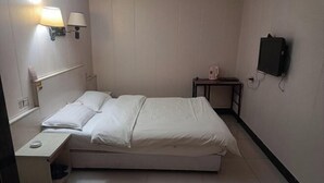 Ein Einzelbett mit weißer Bettwäsche in einem Hotelzimmer. Es gibt einen Nachttisch mit einer Lampe, einen kleinen Tisch mit einem Wasserkocher und einen an der Wand montierten Fernseher.