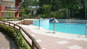 A swimming pool with a wooden deck, a lounge chair, and a building with a balcony in the background.