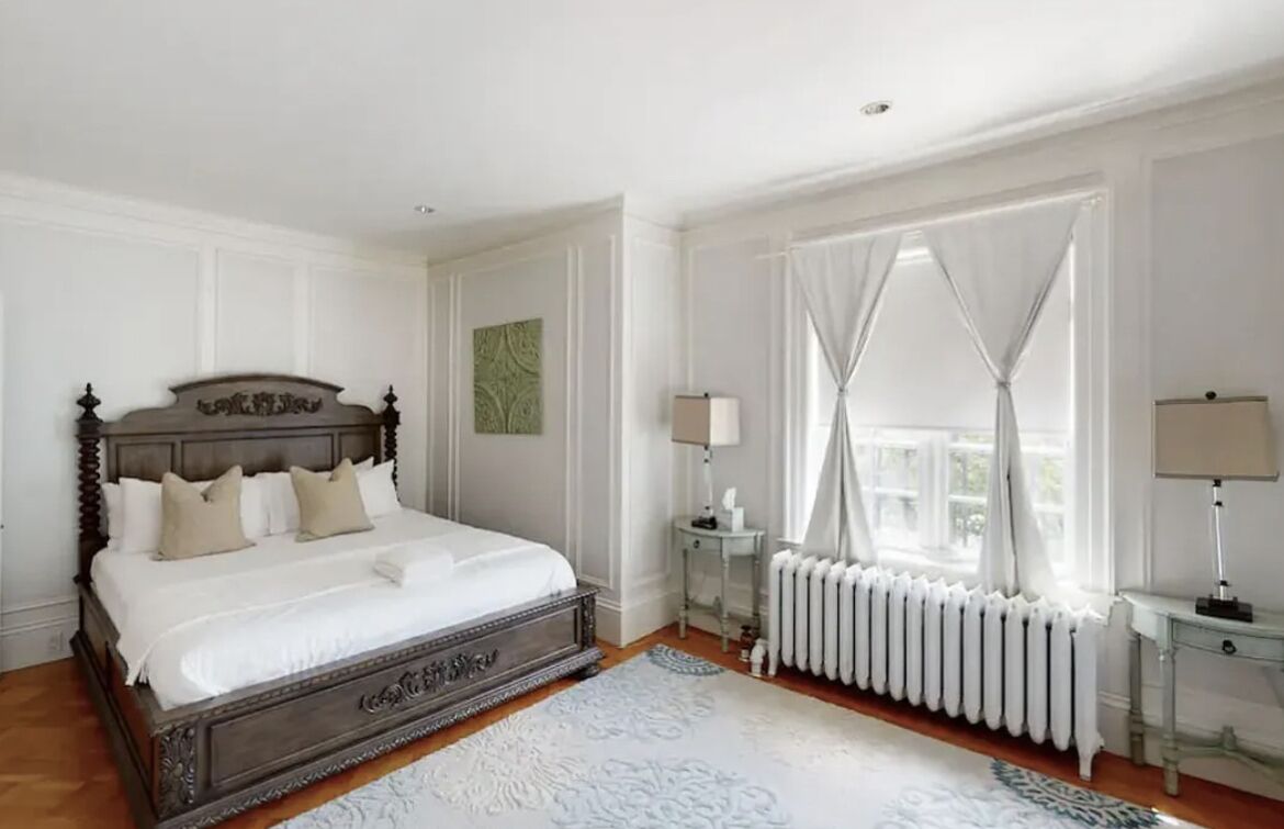 Un dormitorio con una cama grande, paredes blancas, un radiador y una ventana con cortinas.