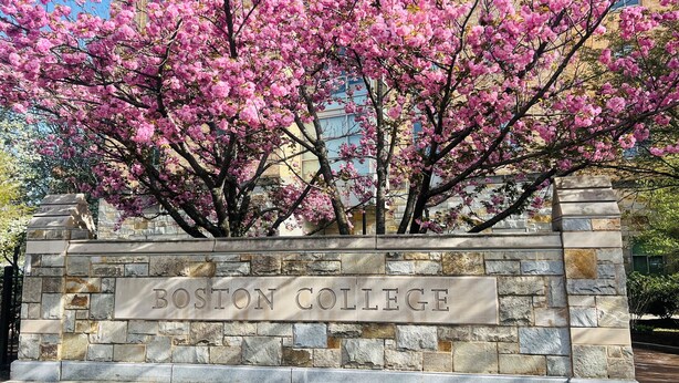 A stone wall with the inscription "BOSTON COLLEGE" and a tree with pink blossoms in front.
