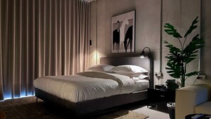 A modern bedroom with a large bed, a headboard, a bedside table, a lamp, a plant, and a window with curtains. - C Living 2 - Executive - 1BD Apartment City View (Riyadh)