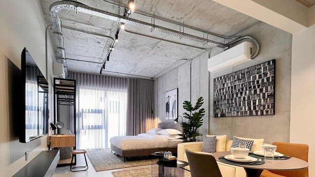 A modern living room with a sofa, coffee table, and dining area. There is a large window with curtains, a flat-screen TV mounted on the wall, and a plant on the floor. - C Living 2 - Executive - 1BD Apartment City View (Riyadh)