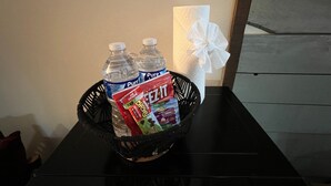Una cesta negra con dos botellas de agua purificada, una bolsa de galletas Cheez-It y una caja de pañuelos blancos sobre una mesa negra.