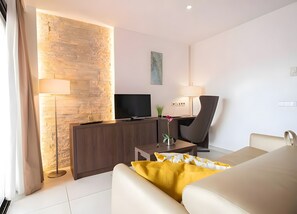 A modern hotel room with a brick accent wall, a flat-screen TV, a desk with a lamp, a comfortable sofa, and a decorative plant. - Apartment 'Suite 3' with Shared Pool, Wi-Fi and Air Conditioning (Cala Agulla)
