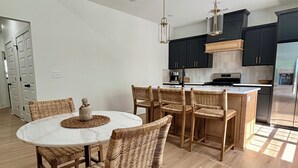 Une cuisine moderne avec des meubles de couleur sombre, un îlot central et un coin repas doté de chaises en osier et d’une table en marbre.