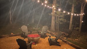 Un espace extérieur aménagé avec des chaises Adirondack et un foyer, entouré d’arbres et illuminé de guirlandes lumineuses.