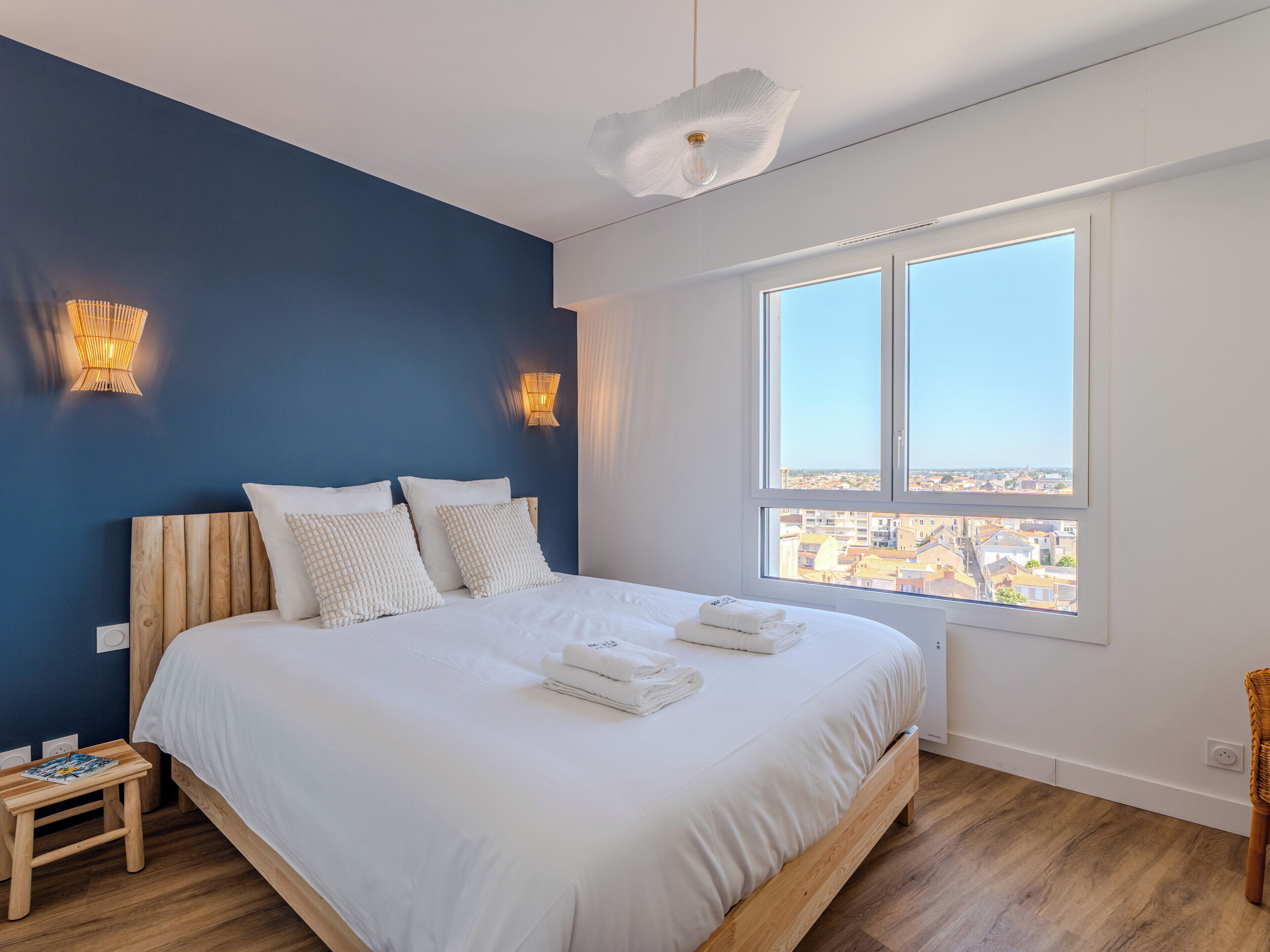 A bedroom with a large bed, two wall-mounted lights, a window with a city view, and a small wooden stool.