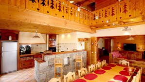 A rustic cabin interior with a stone fireplace, wooden dining table, and a kitchen area. - Apartment 'Alpaga 14' with Private Terrace, Shared Garden and Wi-Fi (Bonneval-sur-Arc)