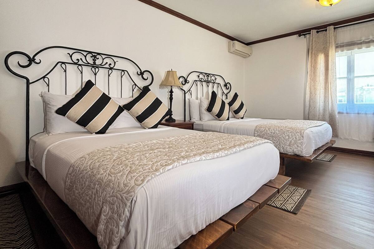 Ein Hotelzimmer mit zwei Betten, Holzboden, einem Fenster mit Vorhängen und einer Deckenleuchte.