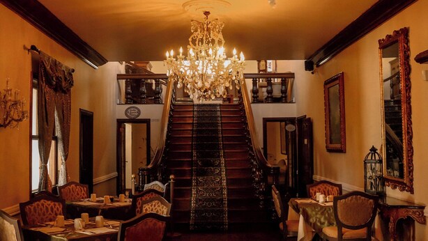 A grand staircase with a chandelier, ornate mirrors, and a dining area with tables and chairs. - Hacienda San Angel (Ensenada)