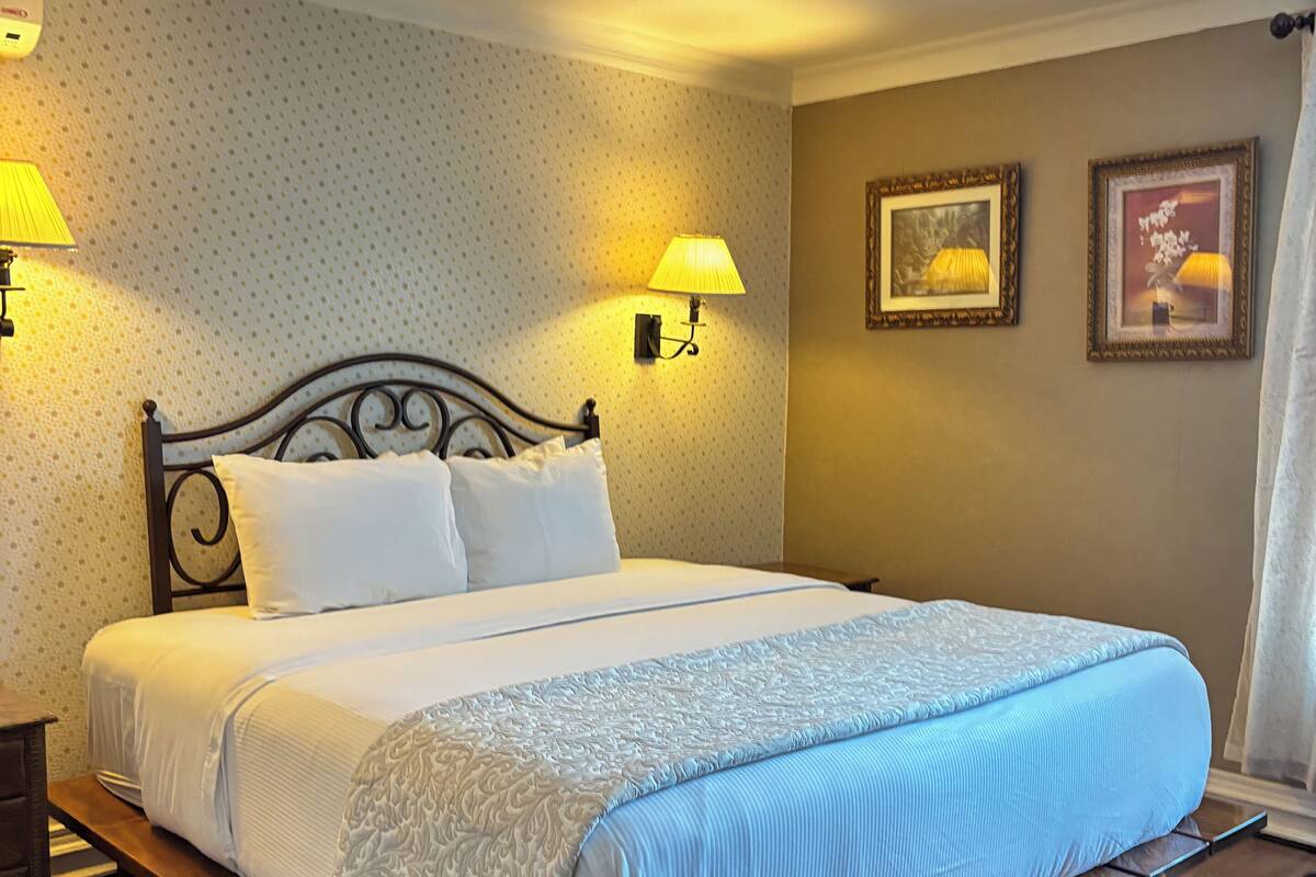 Ein ordentlich bezogenes Bett mit weißblauer Tagesdecke, zwei Wandlampen und zwei gerahmte Bilder an der Wand.