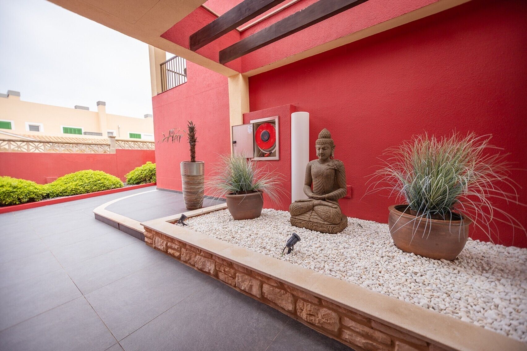 Fachada de un edificio rojo con una estatua de Buda, plantas en macetas y un pequeño jardín.