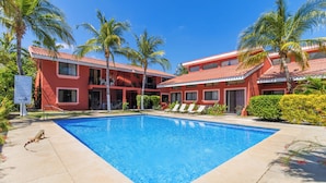 A swimming pool surrounded by palm trees and a red building with multiple balconies. - Las Palmas 1BR+Loft w/ Pool—Walk to Beach & Dining (Coco)