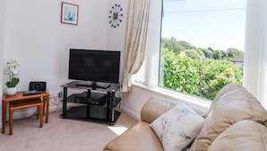 Un salon comprenant une télévision, une horloge, une fenêtre avec des rideaux, un canapé et une petite table sur laquelle est posée une plante.