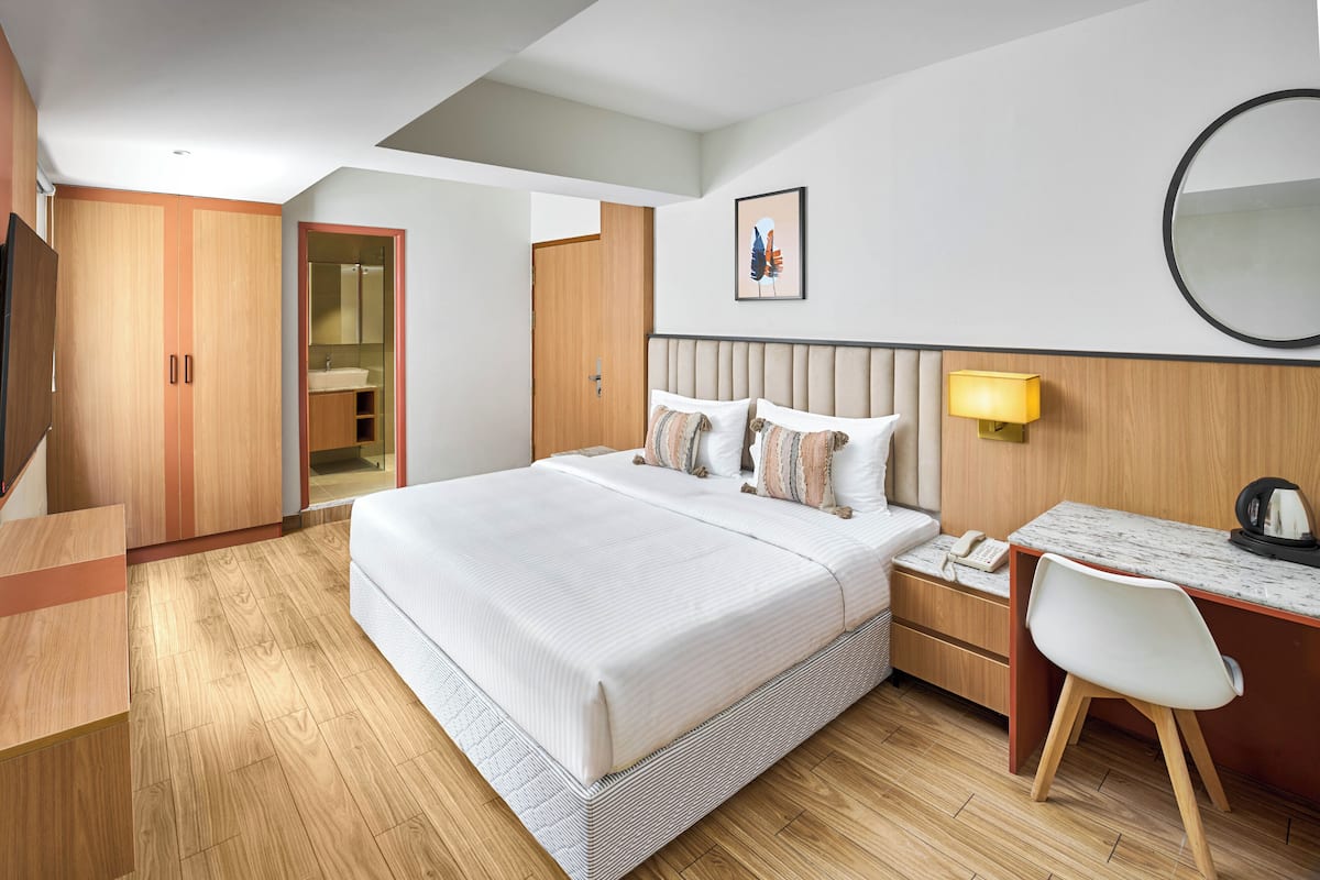 A modern hotel room with a large bed, a desk, a chair, a bedside table, a phone, and a mirror.