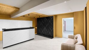 Un espace d’accueil moderne avec un comptoir de réception blanc, un mur en marbre noir et un canapé beige.