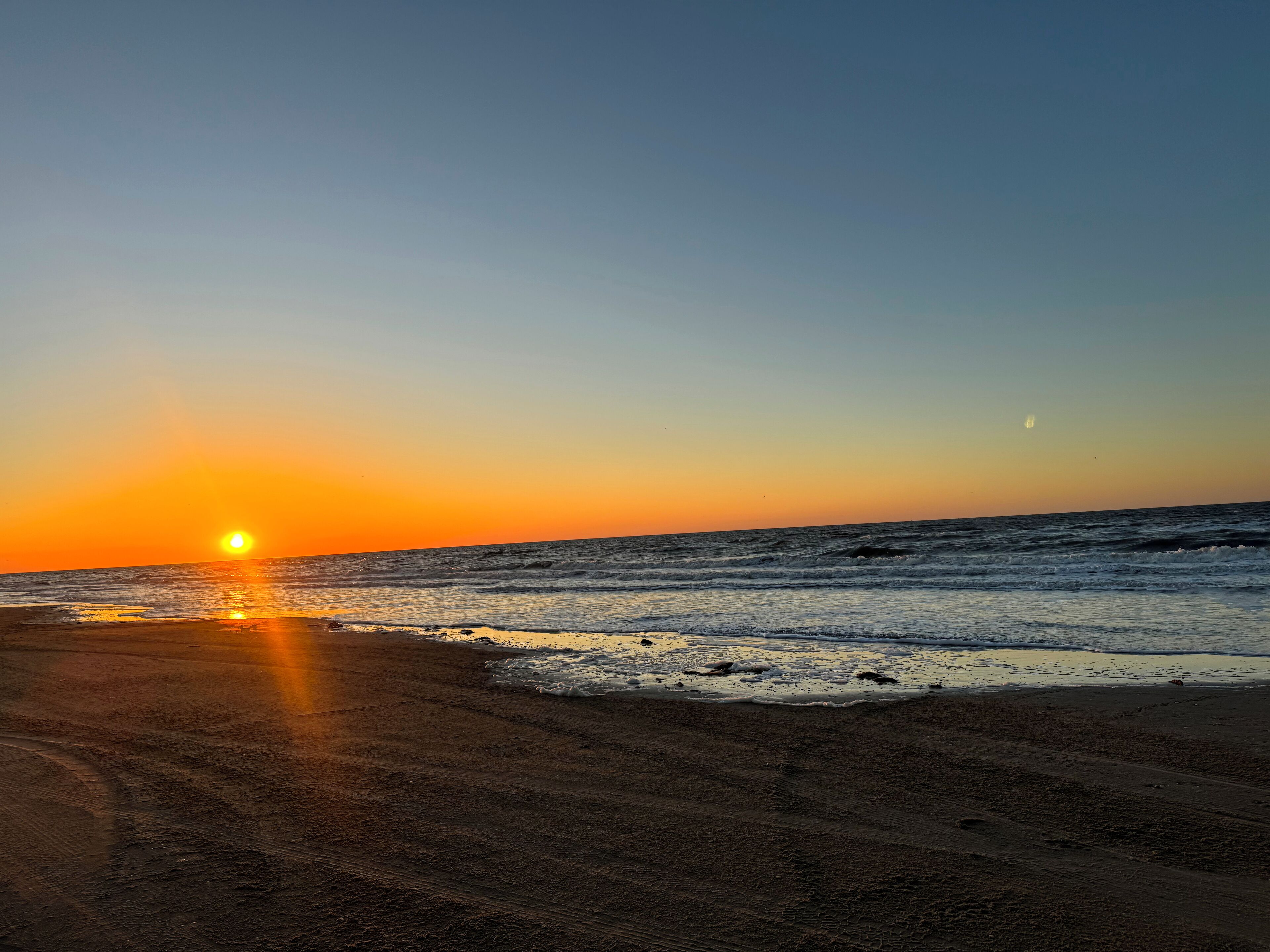 Una playa al atardecer, con el sol bajo en el horizonte y olas suaves lamiendo la orilla.