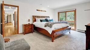 A bedroom with a wooden bed, a nightstand, a lamp, and a view of a forest through a large window. - fishing, hot tub and room to roam! (Shawnee)