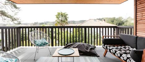 A balcony with a wooden ceiling, a white chair with a patterned cushion, a black sofa with a leaf-patterned cover, and a view of a house and trees in the background.