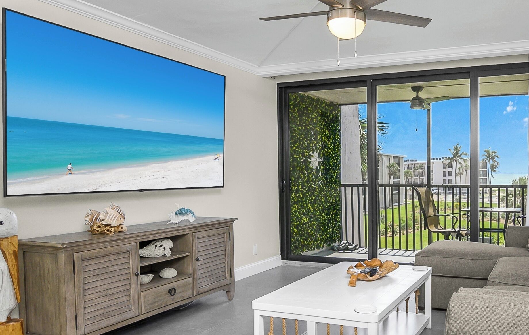 A living room with a large TV displaying a beach scene, a wooden cabinet with decorative items, a white coffee table, and a balcony with a view of palm trees and buildings.