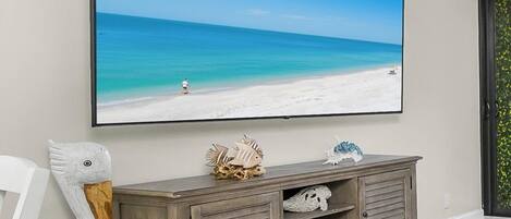 A room with a large framed picture of a beach, a wooden cabinet with decorative items, and a white chair.