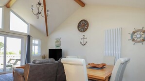 A cozy living room with a sofa, dining table, and chairs. There is a clock, an anchor decoration, and a ship's wheel on the wall. - Lodge 18 (Wadebridge)