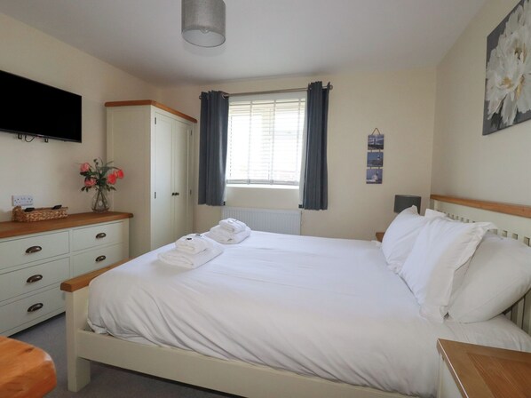 A neatly made bed with white linens and towels, a wooden nightstand, a television mounted on the wall, and a window with curtains. - Lodge 18 (Wadebridge)