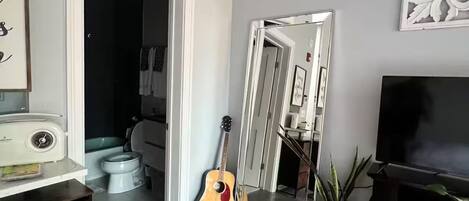 A living room with a guitar, a TV, a mirror, and a staircase leading to a dark room.
