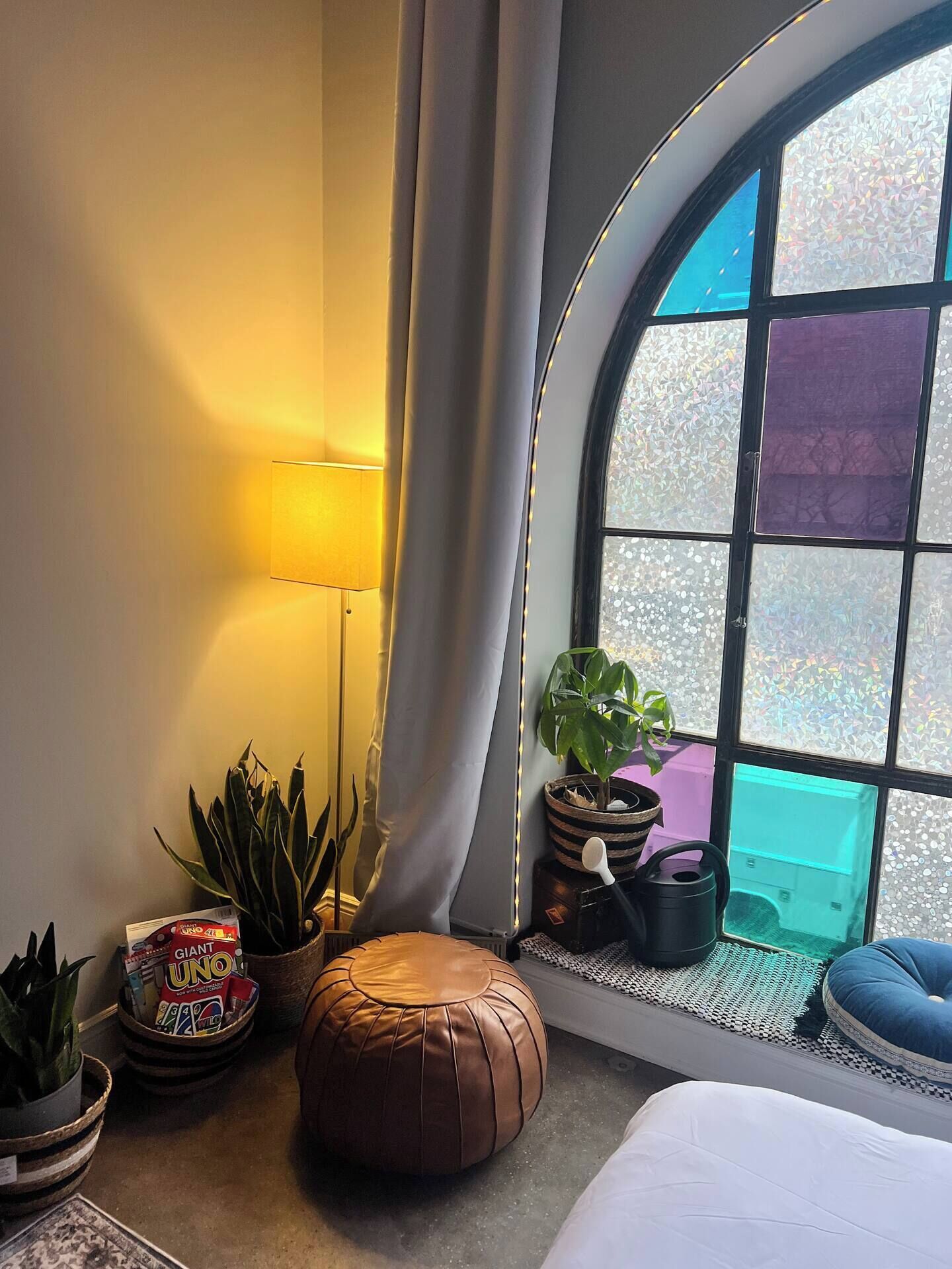 Una habitación acogedora con un ventanal curvo, una lámpara, un sofá y una mesita con plantas y un juego de mesa.