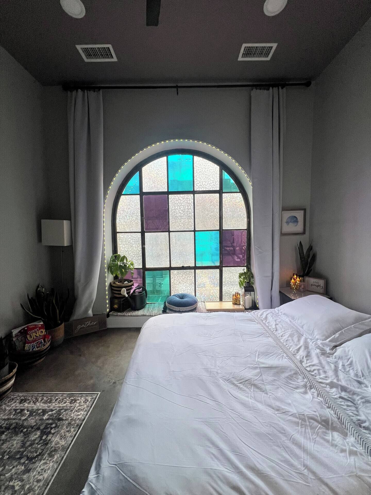 A bedroom with a large arched window, a bed, a bedside table, and a potted plant.