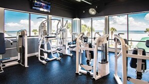 Ein Fitnessstudio mit verschiedenen Trainingsgeräten, großen Fenstern mit Blick auf den Strand und einem an der Wand montierten Fernseher.