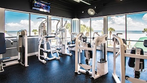 Une salle de sport équipée de divers appareils d’exercice, de grandes fenêtres donnant sur la plage et d’un téléviseur fixé au mur.