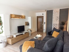 A modern living room with a flat-screen TV, a grey sofa with yellow cushions, a wooden entertainment unit, and a decorative wall piece. - Cozy, bright apartment with fully equipped kitchen and parking space (Bad Berneck)