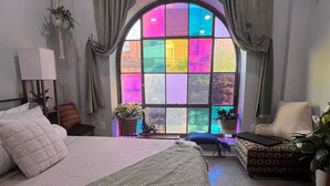 A bedroom with a large bed, a chair, a small table, and a window with colorful glass panes.