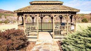 A wooden gazebo with a tiled roof, surrounded by a stone pathway and dense shrubbery.