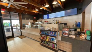 A coffee shop interior with a counter, shelves with snacks, a menu board, and a ceiling fan.