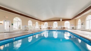 Une vaste piscine intérieure à l’eau d’un bleu limpide, entourée de chaises longues et de tables, et dotée de grandes fenêtres en arc de cercle.