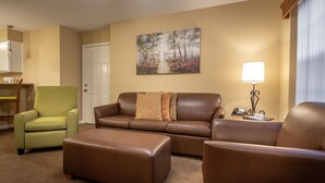A living room with a brown sofa, a green armchair, and a brown ottoman. There is a painting on the wall and a lamp on a side table.