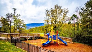 A playground with a slide and climbing structures, surrounded by a wooden fence and trees, with mountains in the background.