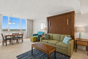 A modern living room with a sofa, armchair, dining table, and chairs. There is a large window providing a view outside.
