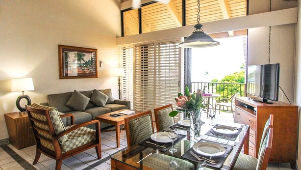 A living room with a sofa, chairs, a coffee table, and a dining area with a table set for two. There is a TV on a stand and a balcony with a view of greenery.