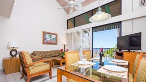 A dining area with a glass table, wooden chairs, and a sofa. There is a TV on the wall and a window with blinds. - 1BR Oceanview Condo w/ Pool & Balcony in Kihei (Kihei)