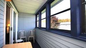 A covered porch with two plastic chairs and a table, large windows with dark trim, and a view of a bridge and trees.