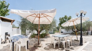 An outdoor patio with stone flooring, white plastic chairs, and tables, shaded by large umbrellas.