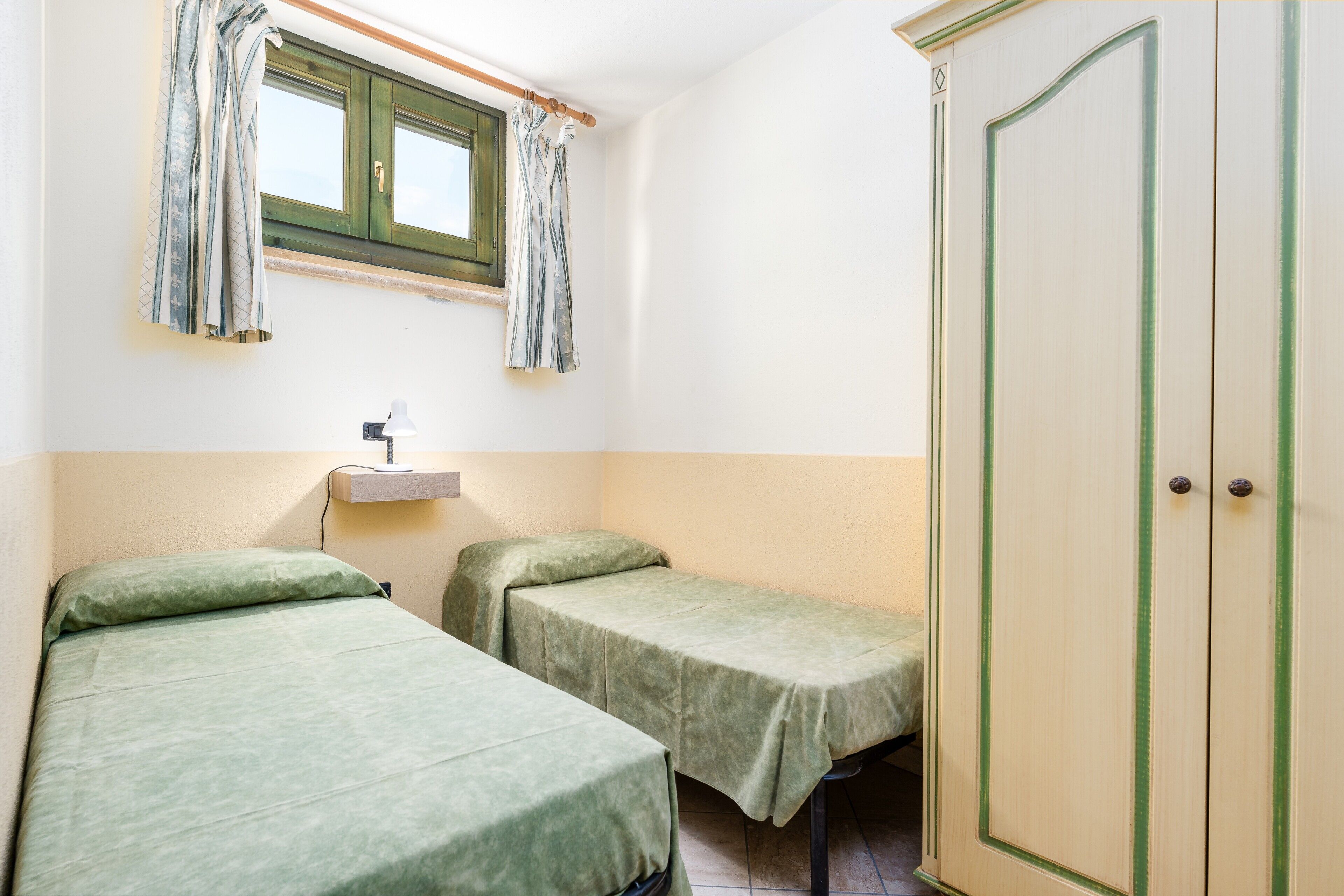 A room with two single beds, a wardrobe, a window with curtains, and a bedside table with a lamp.