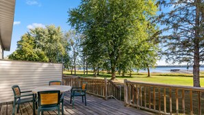 Una terraza de madera con mobiliario exterior, vistas a un lago y un área verde con árboles.