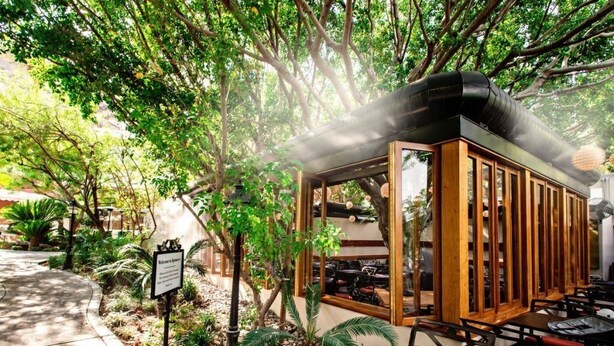 Un espace de restauration en plein air, avec des tables et des chaises en bois, entouré d’une végétation luxuriante et d’arbres.