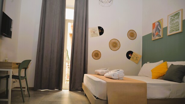 Ein Hotelzimmer mit Bett, Schreibtisch und Stuhl. Vor dem Fenster sind Vorhänge, und die Wand ist mit Kunstwerken mit runden und rechteckigen Formen dekoriert.