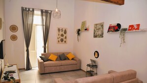 Ein Wohnzimmer mit einer Couch, einem Couchtisch und einem Fernseher. Vorhänge, Wandbilder und eine Regal mit Dekoration sind vorhanden.