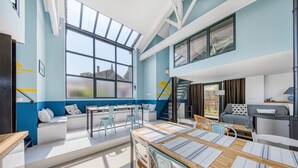 Amplia habitación con un gran ventanal, una escalera, un sofá, una mesa de comedor y una zona de cocina.